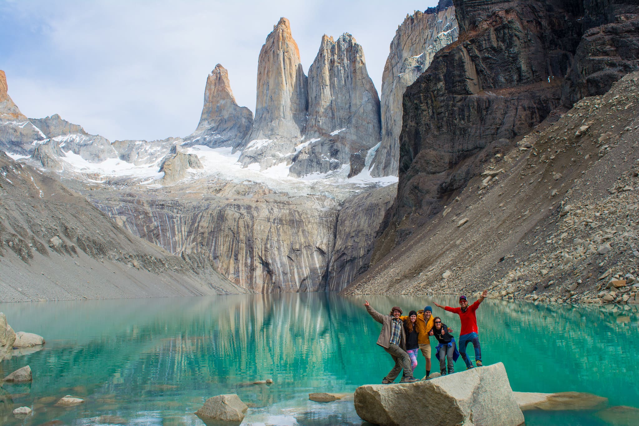 Five hikers posing cheerfully on a rock by a turquoise lake at the base of the iconic Towers in Torres del Paine National Park.