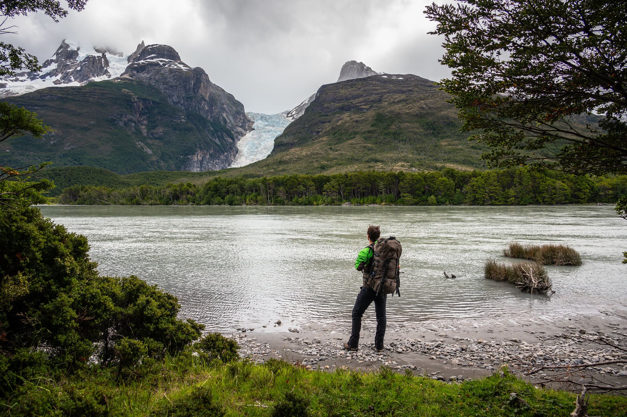 A hiker stands by a serene lake, gazing at a distant glacier surrounded by lush green hills.