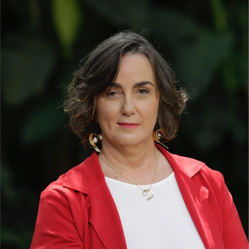 Fotografia de Denise Hills, uma mulher branca, de cabelos castanhos ondulados na altura do pescoço. Ela veste uma blusa branca e um blazer vermelho.