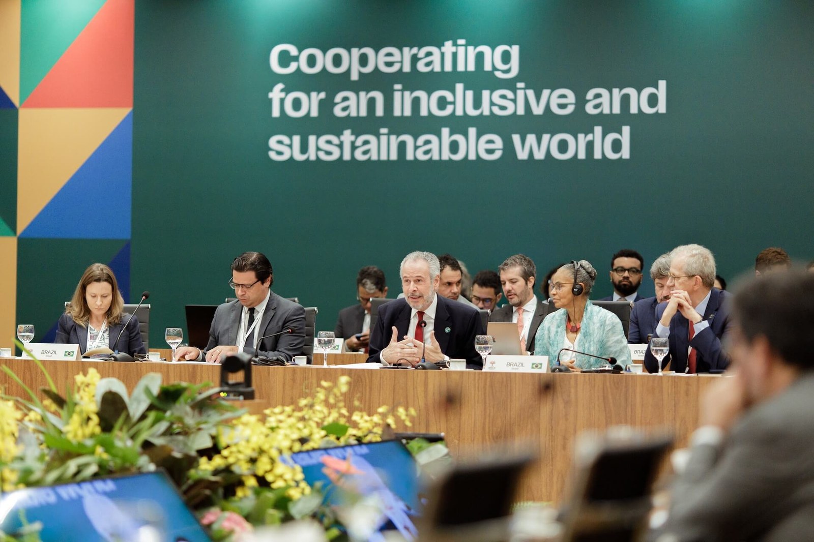 Autoridades reunidas em conferência internacional, sentadas lado a lado em mesa com microfones e crachás; ao fundo, o lema "Cooperating for an inclusive and sustainable world"