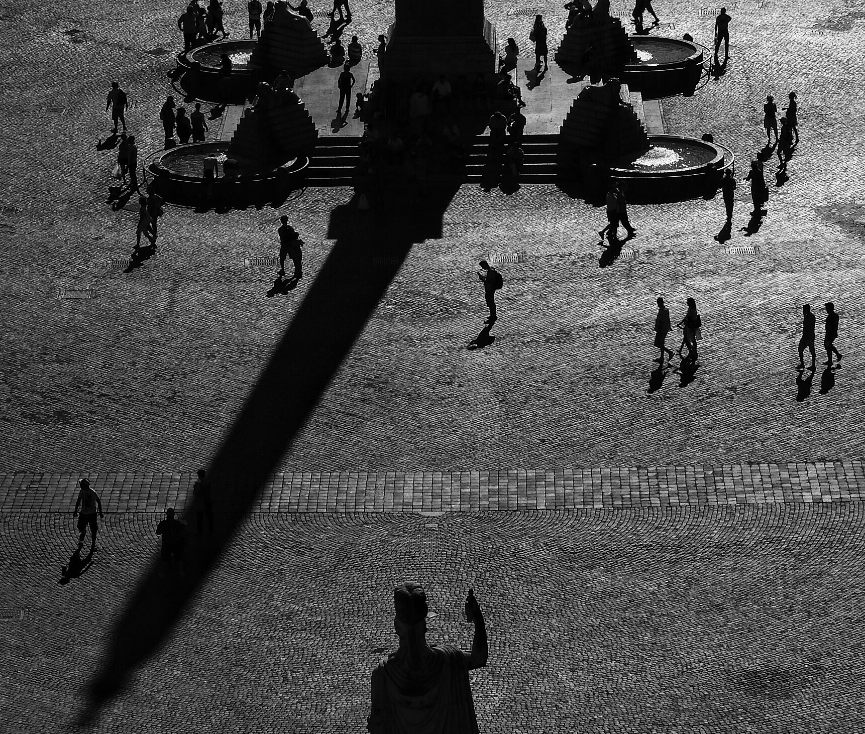 Une place publique vue d'en haut : des passants, l'ombre d'un obélisque, une statue qui lève la main