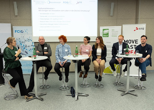 7 Personen sitzen vor einer Leinwand, davor stehen Stehtische.