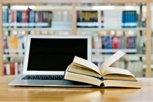Ordinador portàtil i, al costat, dos llibres oberts. En segon pla, la prestatgeria d'una biblioteca