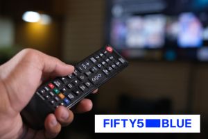 Una mà sosté un comandament a distància. En segon terme, una pantalla de televisió. A la part inferior dreta de la imatge, el logotip de Fifty5Blue