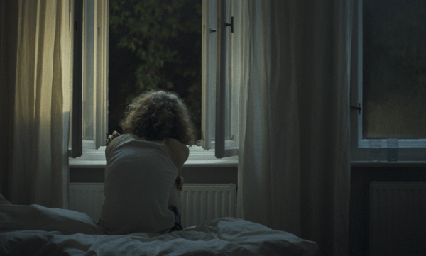 Eine Person sitzt nachts auf einem Bett und blickt durch ein geöffnetes Fenster nach draußen. In einer weiteren Szene steht dieselbe Person mit lockigen Haaren im Freien und schaut zur Seite, umgeben von einem urbanen Umfeld. Danach sitzt sie allein an einem Tisch mit zwei Tellern in einem stillen Innenraum. Es folgt ein dunkler Blick durch schmale, vertikale Öffnungen in einen Raum. Zum Schluss stehen zwei Gläser auf einer Fensterbank, von Tageslicht beleuchtet, wie ein ruhiges Stillleben.