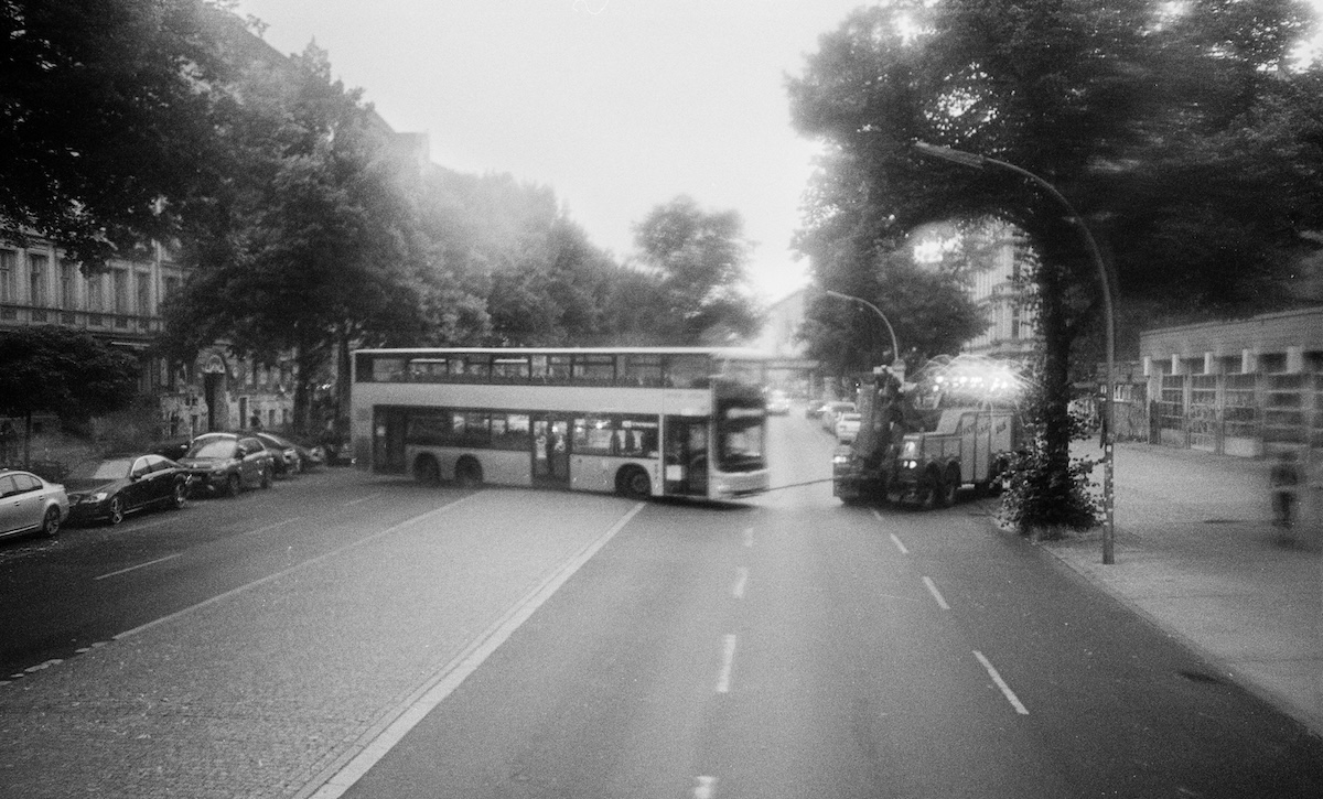 Schwarzweißaufnahme einer breiten Stadtstraße: Ein Doppeldeckerbus fährt quer über die Kreuzung, rechts steht ein Einsatzfahrzeug mit eingeschaltetem Licht. Bäume und Wohnhäuser säumen die Straße, leichte Bewegungsunschärfe erzeugt eine dynamische Stimmung.