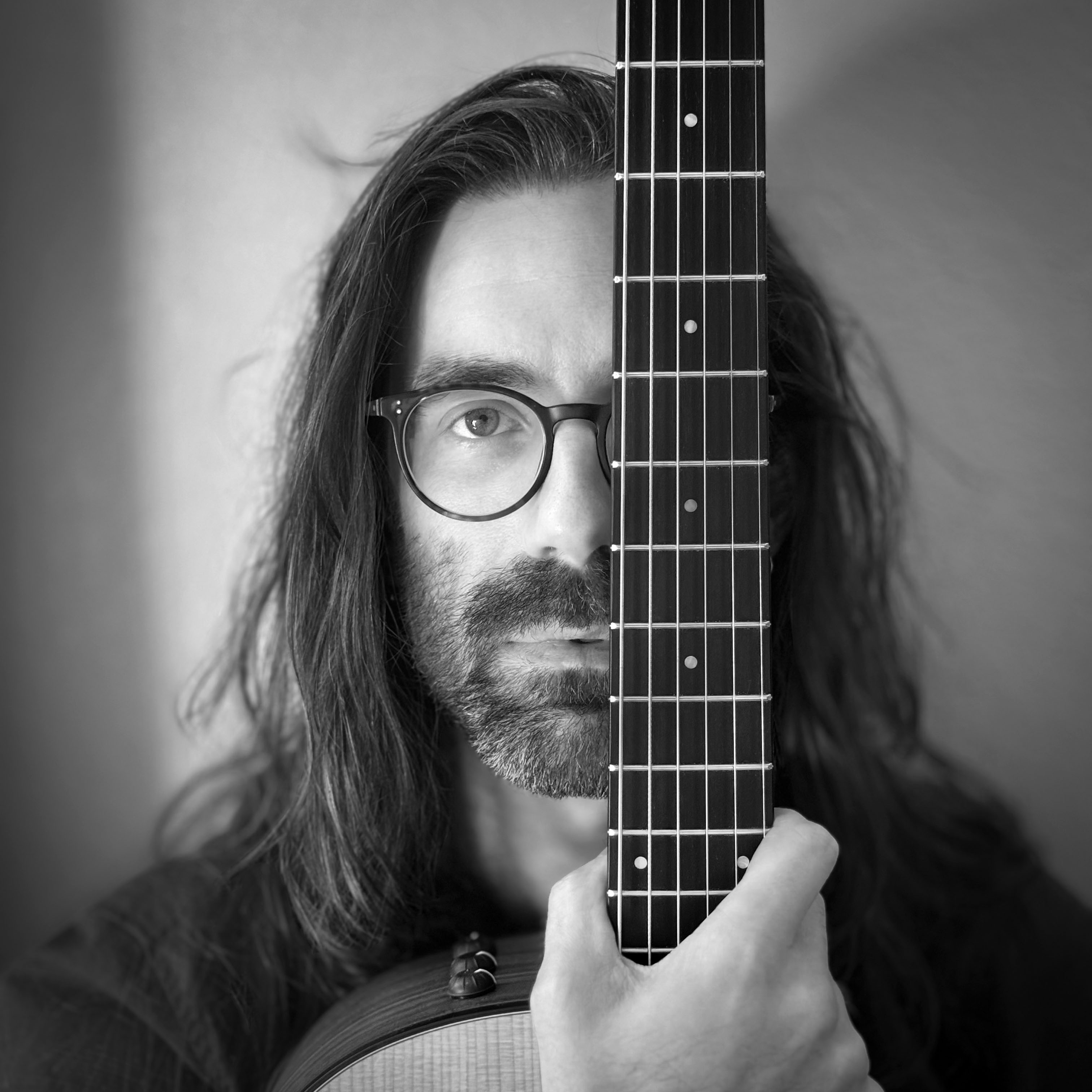 A black-and-white headshot of a man with long hair and a short beard, wearing glasses, looking directly at the viewer while covering his left eye with the fretboard of a guitar held vertically in his right hand.