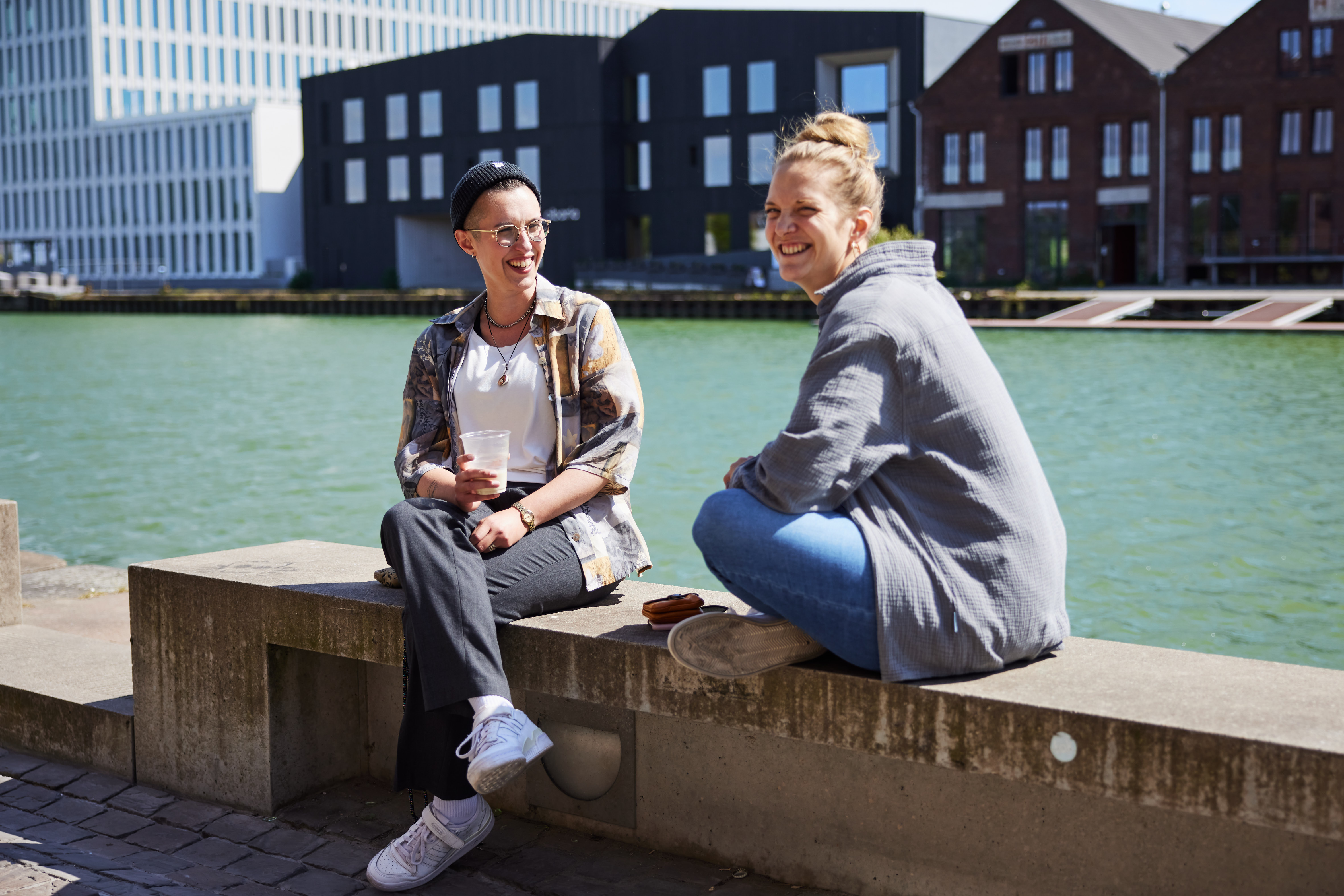 Eli Doudis und Ronja Runge sitzen auf einer Steinmauer. Im Hintergrund der Münsteraner Hafen und dunkle Gebäude. Eli sitzt links, überschlägt die Beine und lächelt Ronja an. Ronja sitzt seitlich zur Kamera im Schneidersitz.