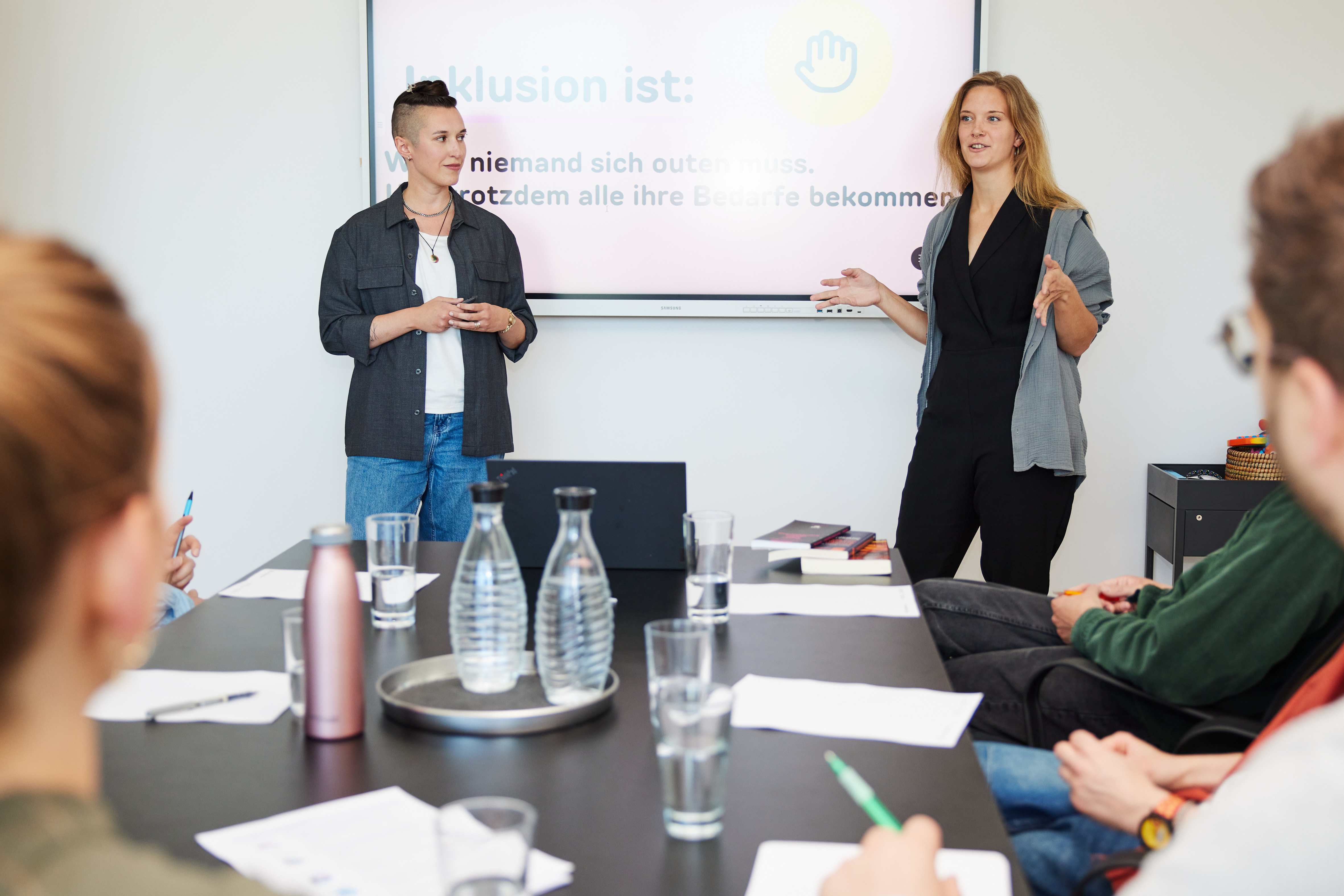 Eli Doudis und Ronja Runge stehen vor einer Leinwand und sprechen in Richtung Kamera. Eli steht links in grauem Blazer. Ronja ist schwarz gekleidet und gestikuliert. Vorn im Bild sind mehrere Menschen von hinten zu sehen.